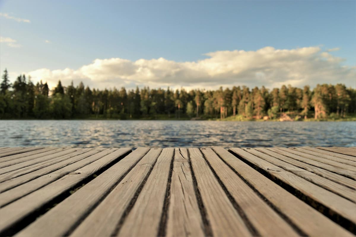 A weathered wooden lake dock stretches toward a calm Swedish lake at golden hour, surrounded by pine forest under a blue summer sky, Sweden