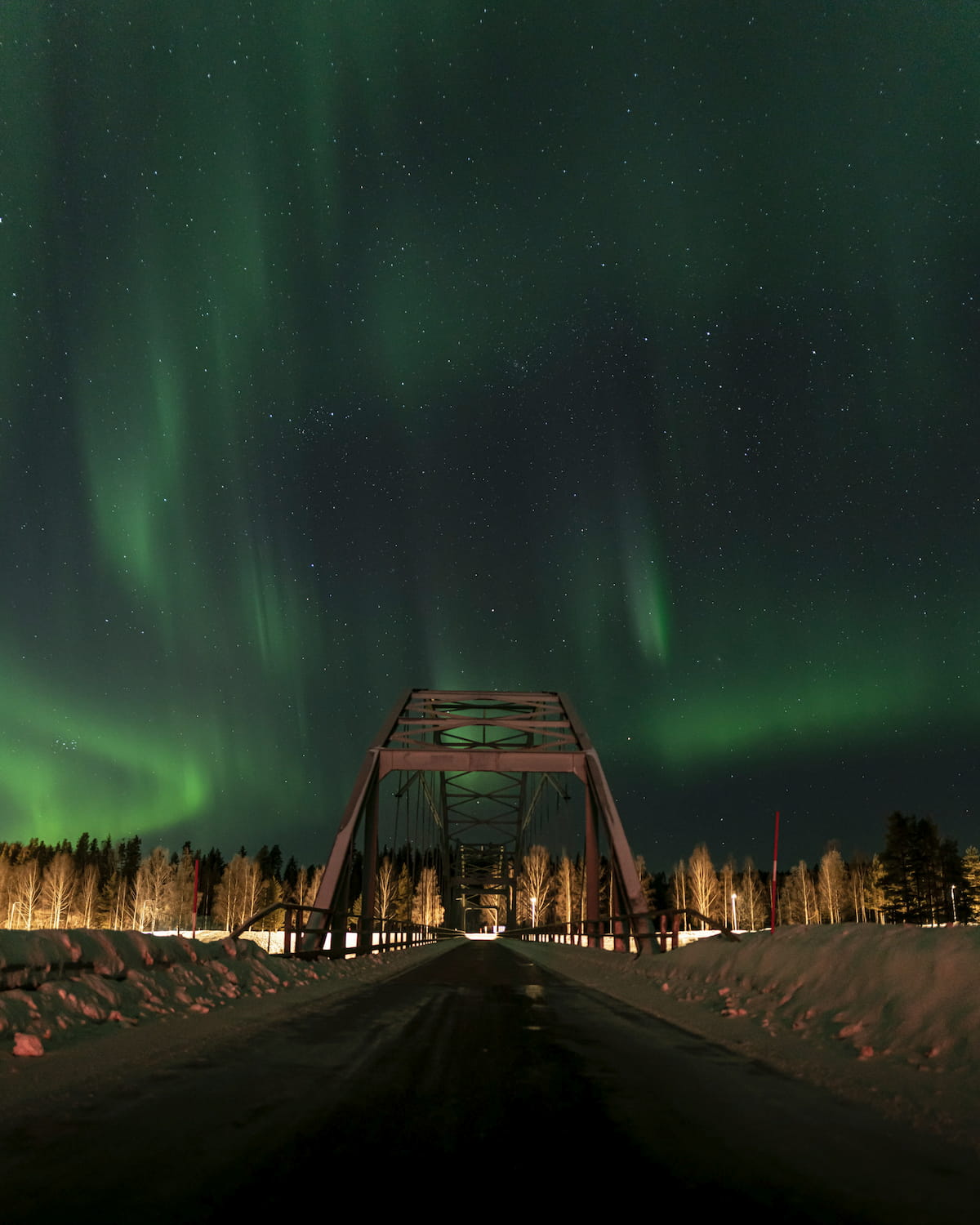 Northern lights dancing over a steel bridge on a snowy Lapland road at night