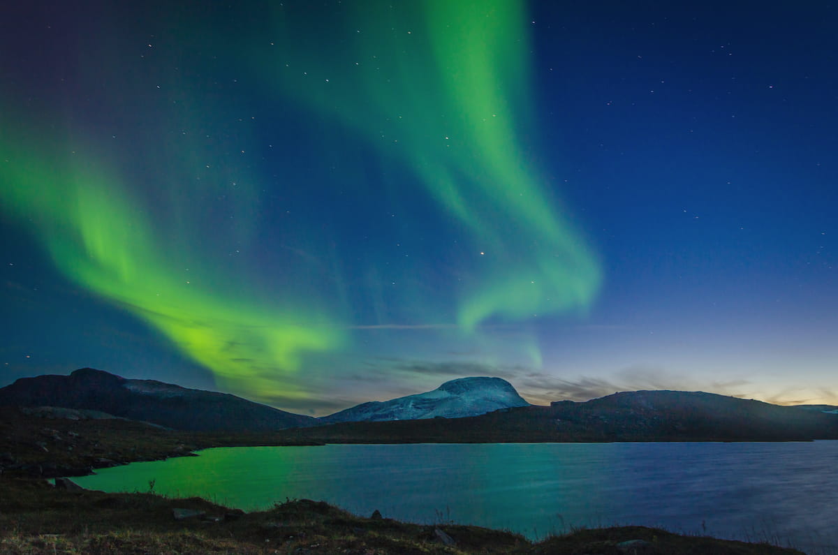 Brilliant green northern lights reflected in a still lake beneath the mountains of Swedish Lapland